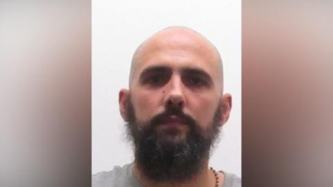 A custody photo of a man looking straight at the camera. He has a full brown beard and he is wearing a grey top and a beaded brown necklace.