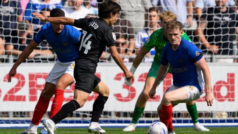 Koki Saito, wearing QPR's black away kit, moments before shooting to score against Portsmouth. The Portsmouth defenders, wearing their blue, white and red home kit can be seen trying to block his effort.
