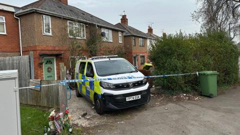 A police van is parked on the drive of a house. Police tape stretches across the drive from a green bin to a fence panel. Some flowers have been laid to the left of the drive, by the fence panel. The front door of the house is green with red brick around the lower level of the house it and brown render above.