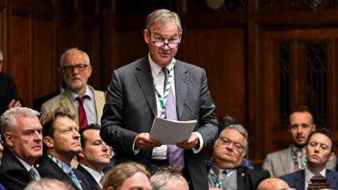 Rupert Lowe stands and speaks in the House of Commons, holding a piece of paper. He wears a suit, tie, and glasses. The chamber is filled with other formally dressed members, with wooden panelling and ornate architectural details visible in the background.