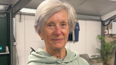 Celia Duff is smiling at the camera and wears a light green hoodie and small gold hooped earrings. She has light grey short hair and a side fringe.