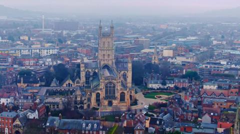 An aerial shot of the centre of Gloucester showing the cathedral in the foreground, and streets fanning out from it. The image is taken on a hazy day