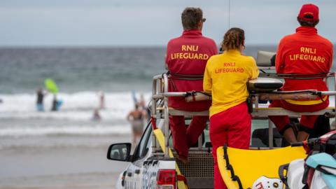 Two male lifeguards are sitting on the roof of a pick-up truck and and one female lifeguard is standing in its flatbed. They are in branded uniform and are looking towards the sea. The sea is blurry as are the swimmers and body boarders in its. 