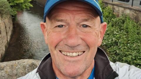 A grinning Eddie Towler sports a blue cap and light grey jacket as he stands on a bridge over a stream