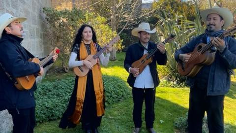 Four members of a band play guitars outside on some grass.