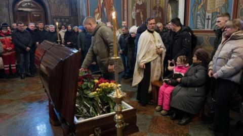A man pays respects to a recent casualty, with the open coffin with flowers in the middle of a church hall, and other mourners all around 