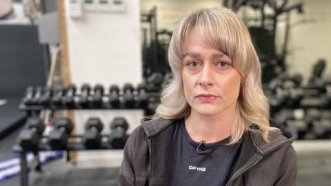 Georgina Cox is sitting in a gym with dumbbells on a rack in front of a large mirror behind her. She is wearing a black t-shirt and a black hoodie over the top of it. She has blonde hair down to her shoulders and looks upset.