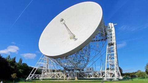 Jodrell Bank radio telescope, a huge white dish with a central tower pointing out of it on a white scaffold, stands in the green field surroundings of the observatory