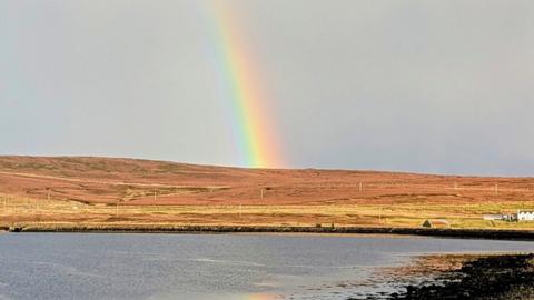 A rainbow arcs out of the centre of fields in shades of brown with grey sky and grey water in the foreground 