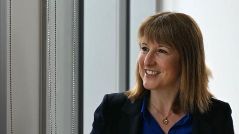 Image shows the Chancellor.
She has bobbed brown hair and is wearing a blue shirt, with a blue jacket and golden necklace. 
She is standing in front of a window looking out of it to her right. 