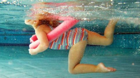 Child in a stripy swim suit with a pink ring around the waist swimming under water. There are lots of bubbles on the water's surface