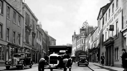 Black and white photograph of old cars driving down a street made of stone buildings.