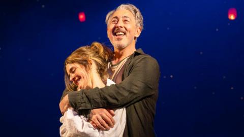 Two performers stand together on a dimly lit stage, framed by a deep blue backdrop and glowing red lanterns suspended overhead.
Warm spotlighting highlights the embrace, while the floating lights create a dreamlike, theatrical atmosphere.