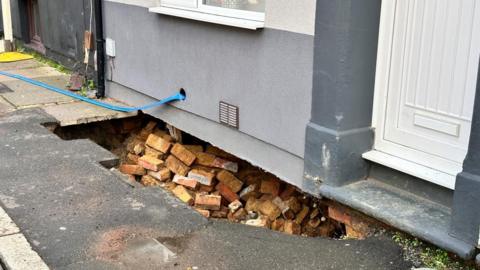 A hole underneath the bottom of a grey coloured house with collapsed red bricks.