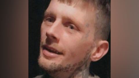 Kyle Pugh had light brown hair with a fringe and short sides. He had facial stubble and tattoos of pawprints along his jaw, trailing up the side of his face. He also had tattoos on his neck.