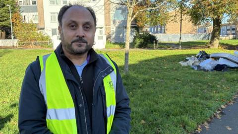 A man with black hair and a beard, wearing a yellow high vis jacket with the 'Bristol City Council' logo, is standing next to an area of grass. In the background is a row of flats and on the grass behind him is a big pile of fly-tipped rubbish - including household waste and a dirty mattress.