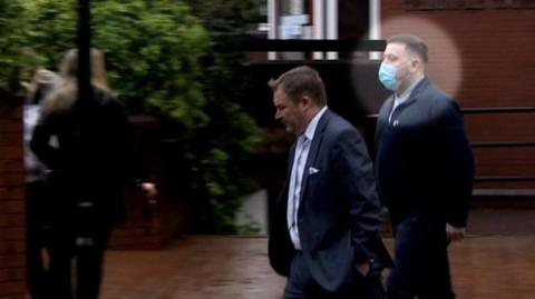 Two men walk along the street outside a court building. One man's face has been digitally illuminated with a circle to identify him as PC Paul Hanson - he is wearing a suit and a blue face mask. He has short brown hair.