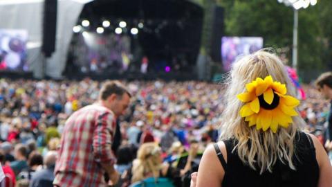 A crowd can be seen in front of a stage. The back of a woman's head is in focus, she is wearing a sunflower hair accessory.