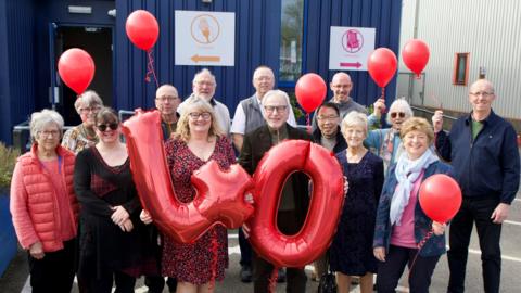 A group of men and women standing together in front of a blue metal-looking building while holding red balloons, A man and a woman in the middle of the group are holding up a "4" and a "0" balloon. They are all smiling and looking into the camera.