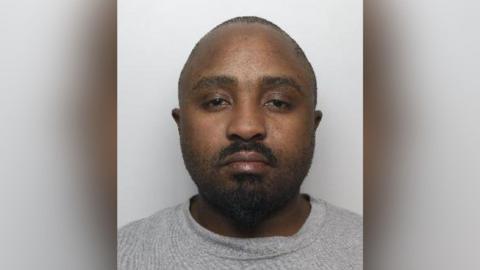 A mugshot of Pfukani, a black man who has a shaven head and a black beard. He is wearing a grey jumper