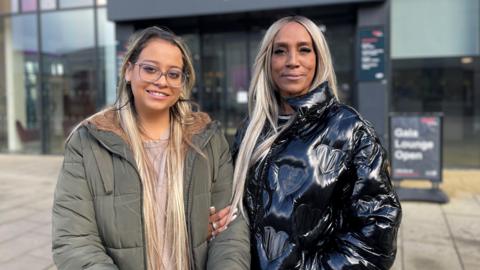 Cheryl Byron, pictured arm-in-arm with her daughter, Abigail. Both women have long, blonde hair and are pictured smiling into the camera standing outside a glass building. Abigail, on the left, wears glasses and a grey coat, her mother stands on the right, wearing a shiny black coat.