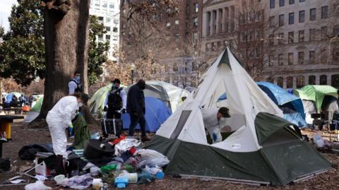 A tent encampment being removed from downtown Washington DC in 2023. Workers in hazmat suits wear face masks. Rubbish is seen strewn outside of one white and green tent. There are large trees and buildings visible outside of the edges of the public park.