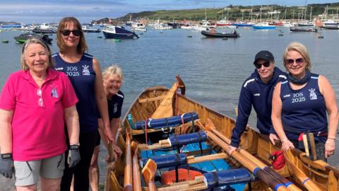 A team of rowers look at the camera while posing with a wooden gig boat in a bay