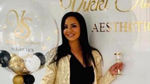 A person stands in a bright, modern interior beside a white wall featuring a gold logo and the text “AESTHETICS.” The person is wearing a black outfit with a gold belt and a gold sequined jacket, holding a large transparent balloon