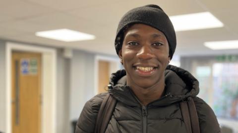 A young black boy, with a coat and hat, standing in large room smiling at the camera