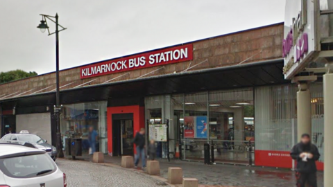 Kilmarnock Bus Station exterior with a red-framed entrance, large sign reading ‘Kilmarnock Bus Station,’ parked cars in front, and people walking near the building.
