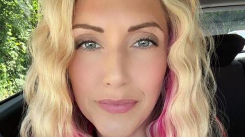 Jessica, who has curly blonde hair and pink highlights, with blue eyes, smiles as she sits in a car. It is a photo of her face. 