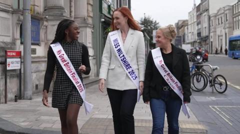 (L-R) Krystel Vida, Alice Cutler and Charlie Cobb, all wearing sashes and smiling are talking and walking towards the camera on a pavement in a built up city