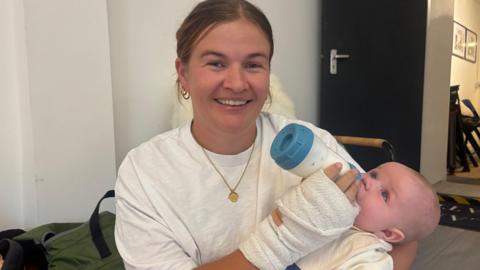 Ceri Jones smiles at the camera holding her baby in her left arm, holding a milk bottle. She has mousy brown hair pulled back in a ponytail and wears a white t-shirt. 