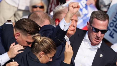 Donald Trump raises his fist after being shot in the ear, which is bleeding, as Secret Service agents move him away.