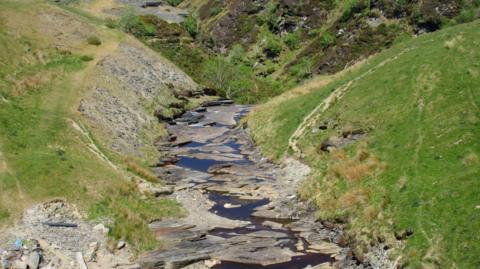 A dry river bed at Rhyd-y-Bedd in Conwy county.  A trickle of brown water is visible, making its way along the rocky river bed.  The grass on the river banks is starting to brown too.
