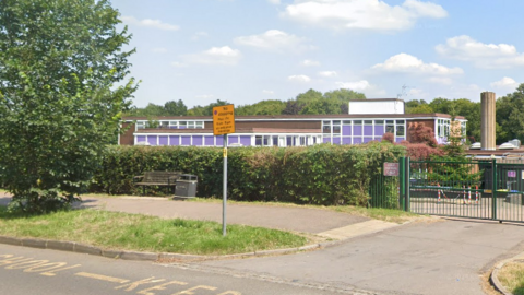 A picture taken next to a road of a brown multi-storey school building with purple windows. There is a bush and green gate guarding the entrance.