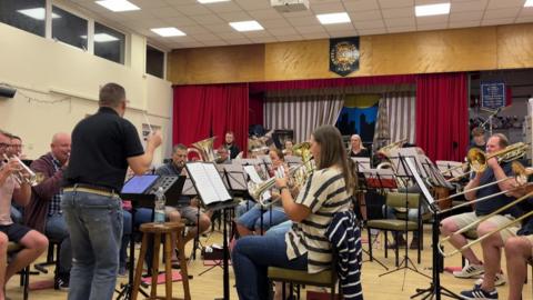 A man in a black polo shirt and jeans conducts a brass band, made up of male and female musicians. There is a trumpet and some tubas in the foreground, and a French horn further back.