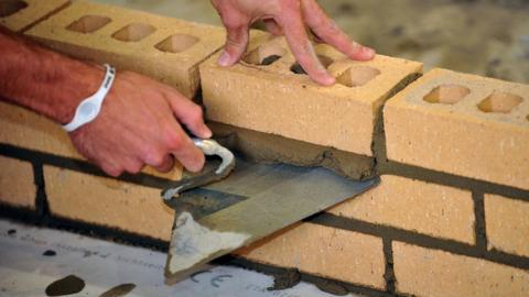A pair of man's hands laying a brick wall with a trowel scraping away spare wet cement.
