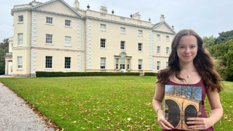 A woman looks towards the camera, stood on a patch of grass in front of a large house. She is holding a book with a photograph on the front. The woman has brunette hair and she is wearing a burgundy top.