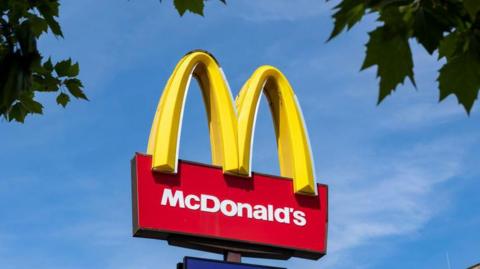 A McDonald's sign against a blue sky with tree leaves at each side 