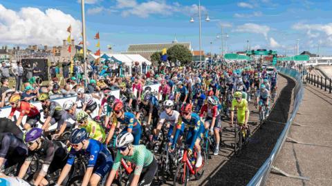 Hundreds of cyclists ride near Lowestoft beach front during the Tour of Britain cycle race. Spectators watch from the left hand side.