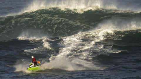 A RNLI lifeguard riding a jetski through a large set of waves. The jet ski is lime green, the lifeguard is wearing a white safety helmet and a red life jacket. He appears small compared to the size of the waves.