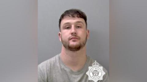 A mugshot of a man with brown beard and fringe. The picture is against a grey background and the man has a serious expression.