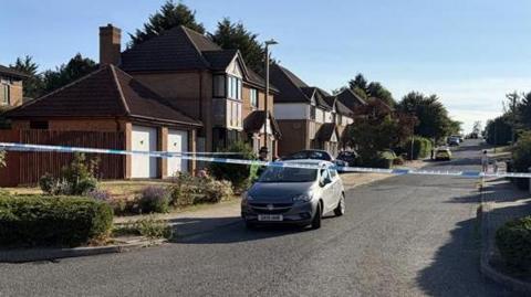 A street in a housing estate with houses on both sides. There is a line of blue and white police tape stretched across the road and a car immediately behind it on the left.