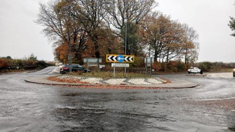 A roundabout on a motorway on a rainy day with grey clouds. A black car is in the process of going around the roundabout. 