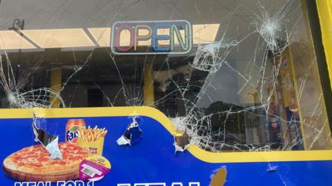 A smashed window of a fast food shop, printed on it is a multi-coloured 'OPEN' sign and a picture of a pizza, fanta can and chips. 
