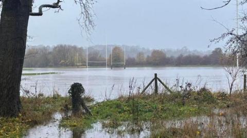 The rugby pitch and surrounding fields are largely submerged in flood water.