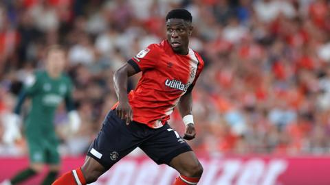 Chiedozie Ogbene looks to his right as he runs on the half turn with his arms by his side while wearing Luton's orange shirt. In the background are blurred fans in the stand