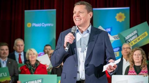 Rhun ap Iorwerth speaking at a Plaid Cymru event. He is smiling and holding a microphone in his hands, wearing a light blue shirt and dark blue jacket. Party activists are standing behind him holding banners.