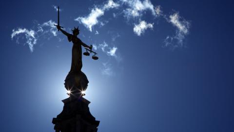 The scales of justice statue above the Old Bailey. The sun rises behind it. There is a blue sky and a few clouds.